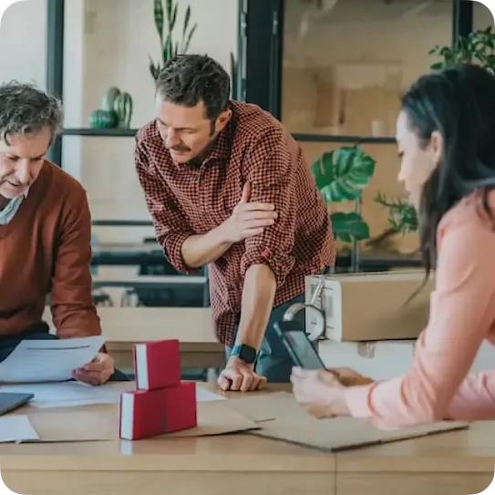 Two men review a document together at a wooden table while a woman holds a smartphone nearby; cardboard boxes and packing tape sit on the table.