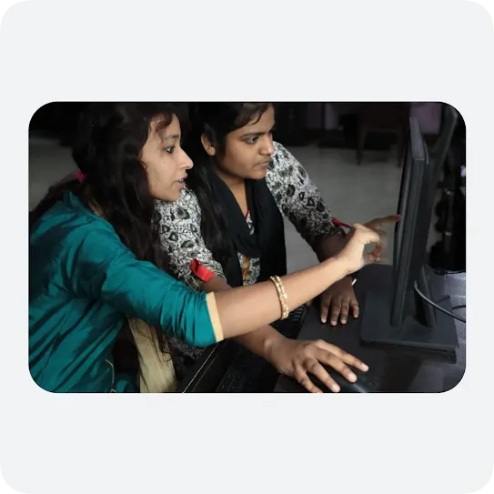 Two female students working together and looking at a computer screen in a classroom setting. The woman in the foreground, wearing a teal top, uses a computer mouse and points toward the monitor screen while explaining something to her peer sitting next to her.