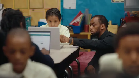 A male teacher in a black shirt leans over a classroom desk to assist a young student using a laptop. They are using the Quill AI-powered app to practice reading and writing comprehension as part of the Policy Hub AI for Society series.