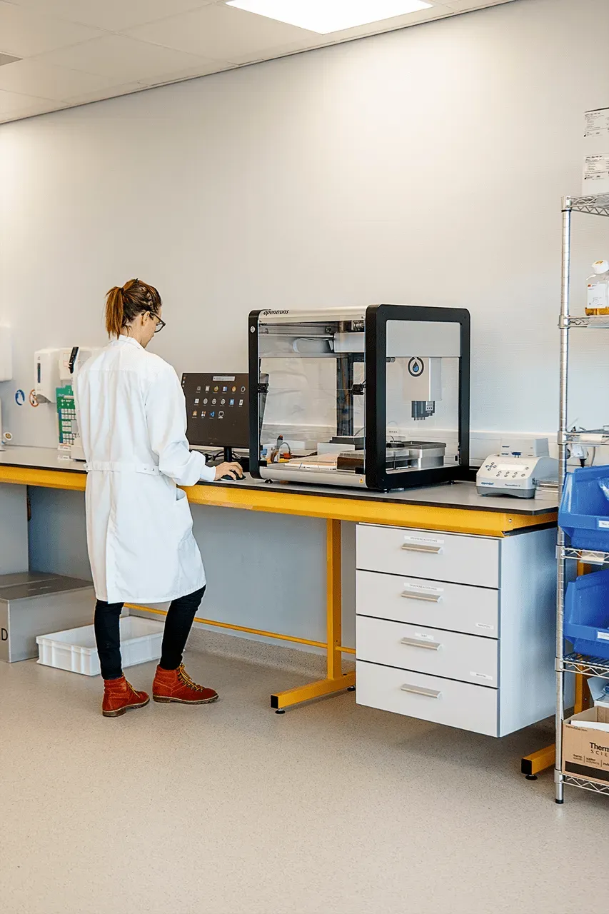 A scientist in a white lab coat and brown boots stands at a laboratory workbench, operating an Opentrons automated liquid handling robot via a computer monitor. This visual represents Cradle’s use of AI to accelerate protein engineering.