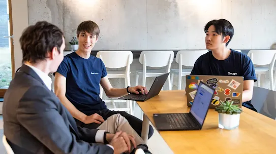 Three young men sit around a wooden table in a modern, sunlit office with a concrete wall and white chairs. One man, wearing a grey suit jacket, sits with his back to the camera, facing the other two. In the center, a man in a navy "One Munich" t-shirt smiles while typing on a laptop. To the right, a man in a navy "Google" t-shirt looks toward the man in the suit; his laptop is covered in colorful stickers. A small succulent sits on the table in the foreground.