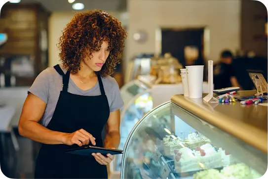 A café worker in an apron prepares an order at a dessert display counter.