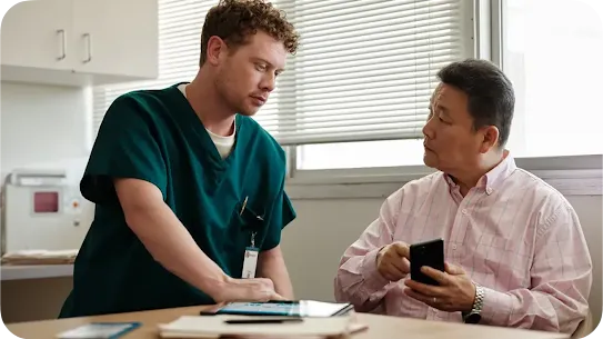 A male healthcare professional in green scrubs leans over a desk to consult with an older Asian male patient holding a smartphone. A tablet and medical documents are visible on the desk in a bright clinic setting.