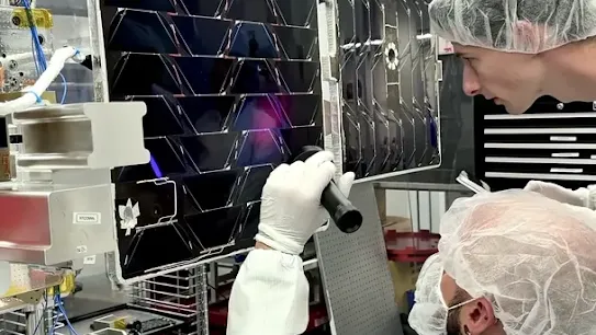 Two technicians wearing hairnets and lab coats inspect large solar panels in a cleanroom lab environment using a flashlight