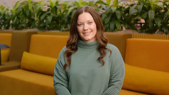A smiling woman with long brown hair wearing a green ribbed turtleneck sweater in a modern, yellow-themed office space.