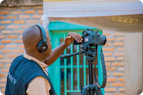 A close-up profile shot of a videographer wearing a "PAGIS TV" vest and large headphones, adjusting the settings on a Canon DSLR camera. The camera is mounted on a tripod under a white canopy, with a brick wall and a teal window frame in the blurred background.