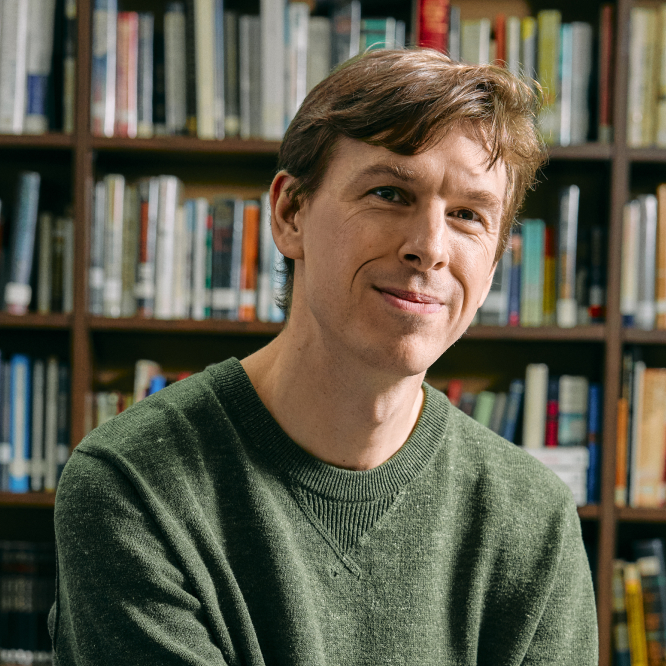 A man with brown hair wearing a green sweater smiling at the camera