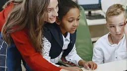 A young female teacher in a red sweater leans over a desk to guide a young girl and boy as they use a computer mouse and keyboard in a classroom setting.