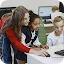 A young female teacher in a red sweater leans over a desk to guide a young girl and boy as they use a computer mouse and keyboard in a classroom setting.