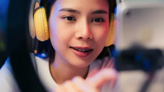 A smiling young Asian woman wearing yellow over-ear headphones while interacting with a smartphone mounted in front of a ring light.