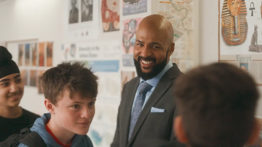 A bald Black man with a beard, identified as Leon Smith, wearing a professional grey suit and blue tie, smiles broadly while looking at students in a classroom. Behind him, the wall is decorated with educational posters, including one about Ancient Egypt.
