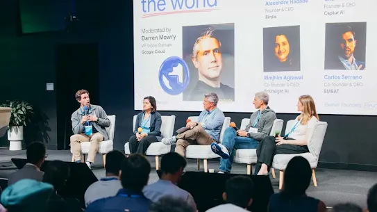A panel discussion taking place on an event stage in front of a live audience. Five speakers are seated in a row on light-colored armchairs. On the far left, a man holds a microphone and speaks to the other panel members—two women and two men—who are listening intently. Behind them, a large projector screen displays speaker profiles, names, and titles under the heading "Moderated by Darren Mowry, VP, Global Startups, Google Cloud" alongside the "Gemini Founders Forum" event branding.