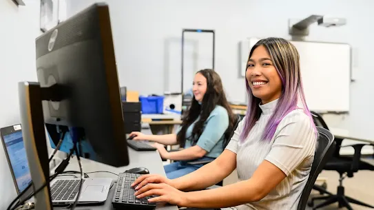 A young woman with purple-tinted hair smiles while practicing digital skills on a computer at a Goodwill training center. In the background, another student works at a terminal, highlighting the Google.org funded workforce development program.