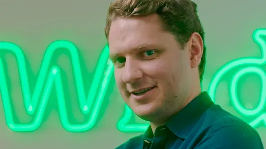 A smiling man in a dark teal collared shirt standing in front of a glowing green neon sign.