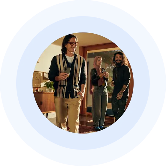 A man holding a smartphone stands with two other people in a living room, all interacting and smiling.