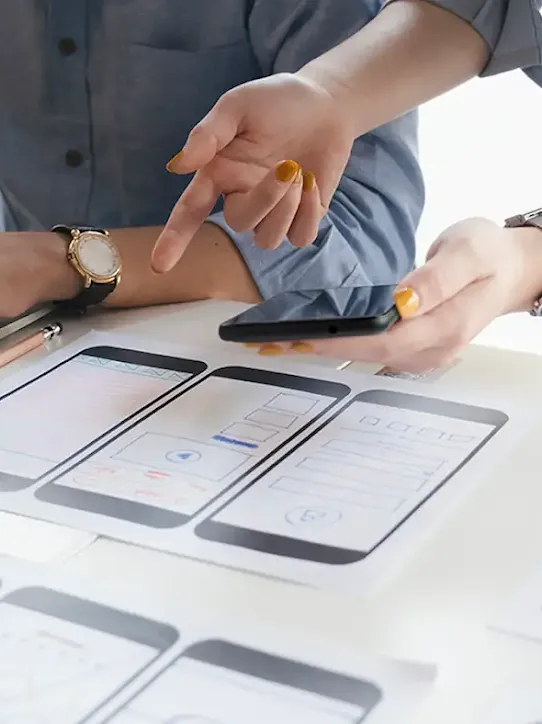 Homem e mulher trabalhando com um notebook e um smartphone. Diversas impressões em cima da mesa mostram mock-ups de telas de celulares.