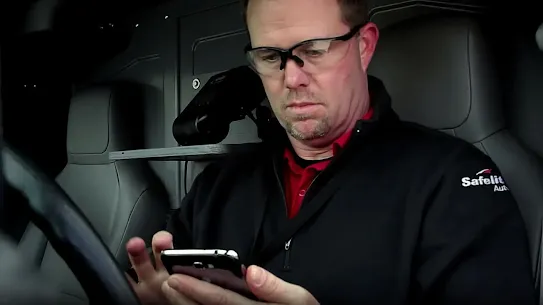 A Safelite AutoGlass technician wearing safety glasses and a company jacket while using a smartphone inside a service vehicle.