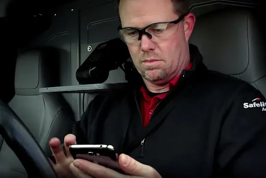 A Safelite AutoGlass technician wearing safety glasses and a company jacket while using a smartphone inside a service vehicle.