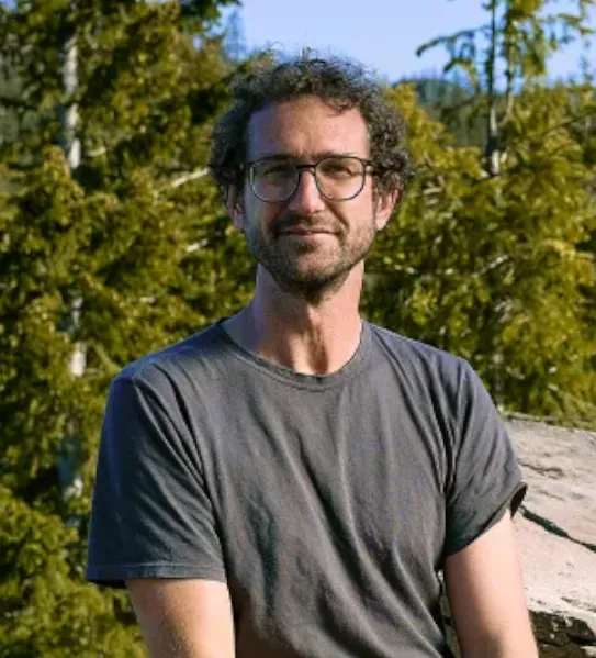 A portrait of Matt Weldy, a wildlife researcher, smiling with glasses and a gray t-shirt, standing in front of evergreen trees in an Oregon forest.