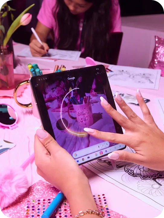 Close-up of hands using a stylus to circle a pink "That's Hot" mug on a folding phone screen, surrounded by colorful craft supplies.