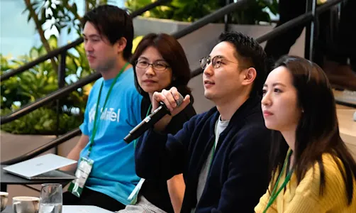 Among four event hosts, one is speaking through a microphone and addressing audience questions.