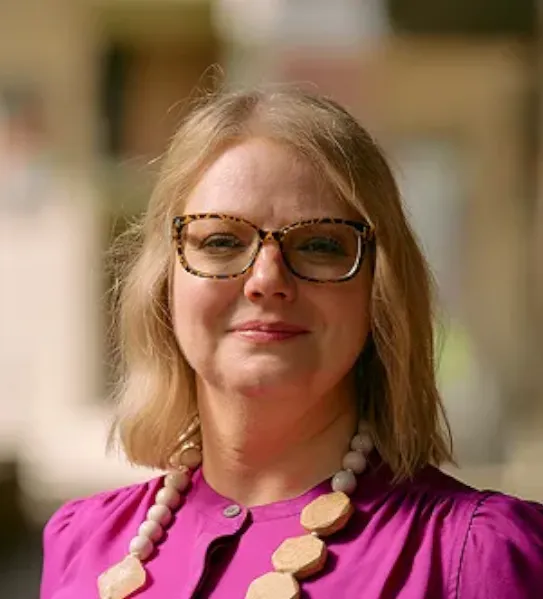 A portrait of a smiling woman with blonde hair and tortoiseshell glasses, wearing a vibrant purple blouse and a chunky beaded necklace, set against a softly blurred outdoor background.