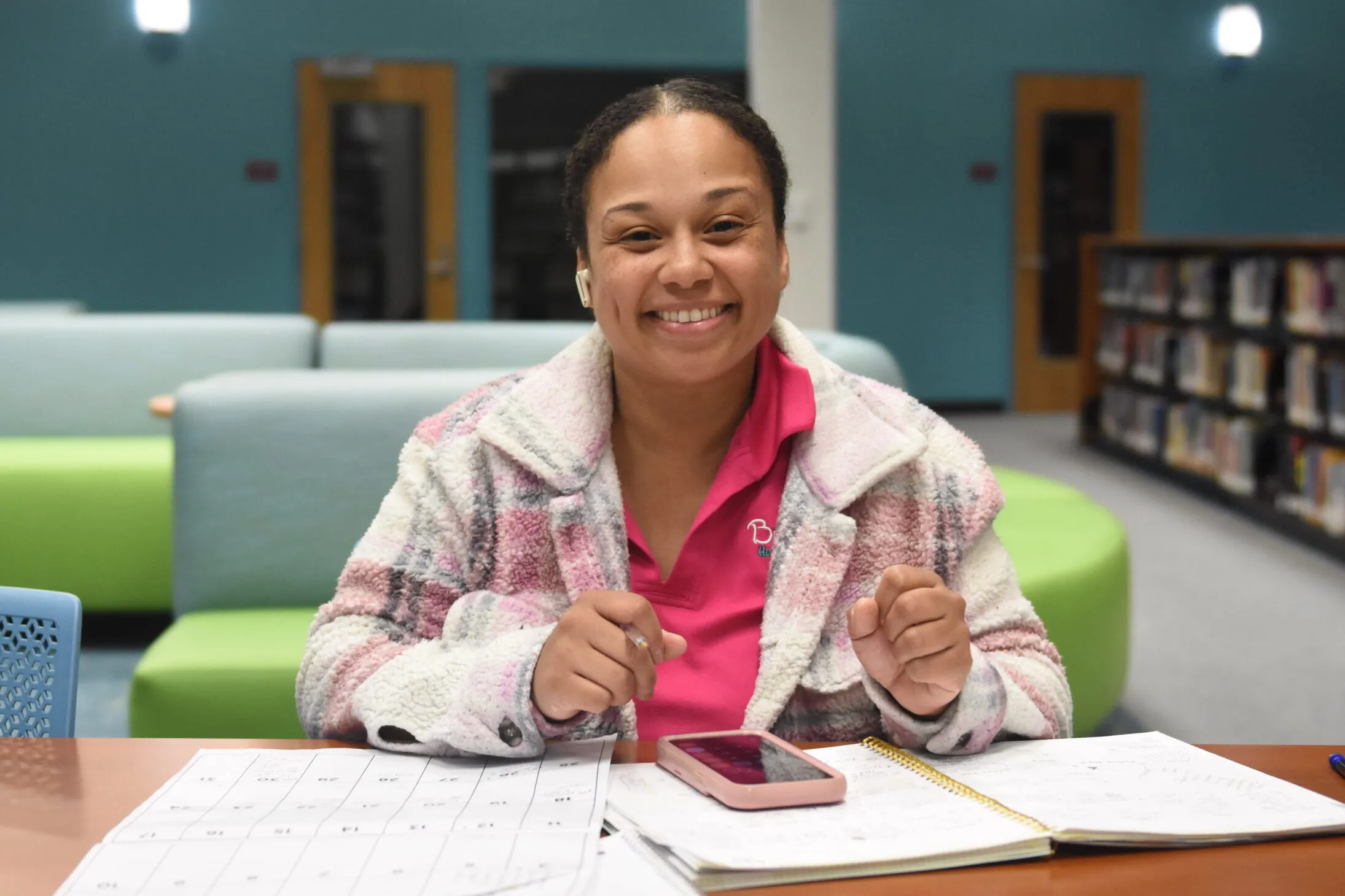 a woman sits at a desk with papers at the library. She smiles at the camera.