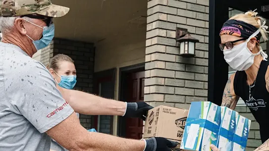 Volunteers wearing protective face masks and gloves distribute boxes of supplies and stacks of medical masks outside a brick building.