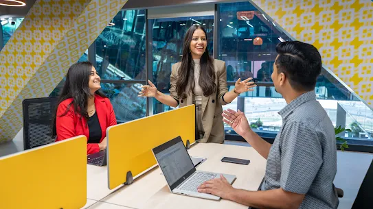 Three coworkers smile and discuss a project around a desk with laptops in a modern, brightly lit office.
