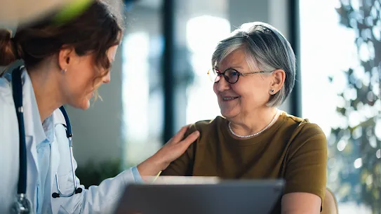A female doctor speaks with a woman with short grey hair and black glasses in an office setting 