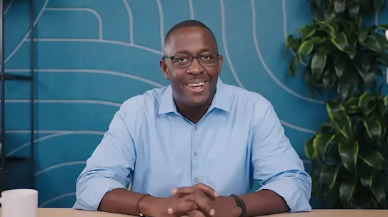 The image depicts a smiling man with glasses sitting at a light-colored wooden table. He is wearing a light blue button-down shirt and has his hands clasped together on the table