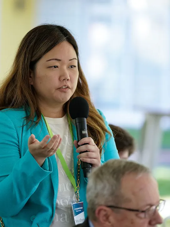 A woman in a vibrant turquoise blazer speaks into a microphone during a Q&A session at the Aspen Security Forum. She is wearing a green lanyard with an identification badge and gesturing with her hand while addressing the audience. The background is softly blurred, showing a bright, airy indoor-outdoor event space.