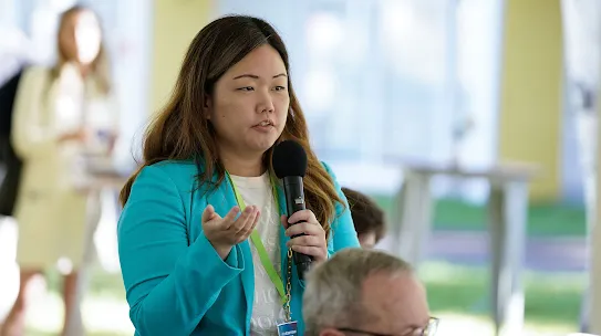 A woman in a vibrant turquoise blazer speaks into a microphone during a Q&A session at the Aspen Security Forum. She is wearing a green lanyard with an identification badge and gesturing with her hand while addressing the audience. The background is softly blurred, showing a bright, airy indoor-outdoor event space.