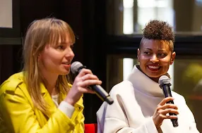A woman is talking on mic and other woman is looking at her while holding the mic.