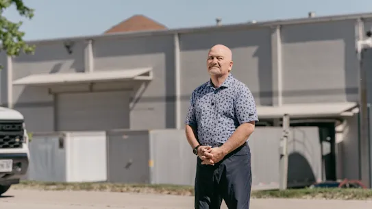 The president of a non-profit that feeds families in need, stands outside an industrial building with his hands clasped and a smile on his face.