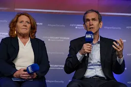 A woman with red hair in a dark blazer and a man in a dark suit speaking on a panel at the Aspen Security Forum. The man is holding a microphone and gesturing while speaking, while the woman sits beside him holding her own microphone. The background is a blue wall with the Aspen Institute and Aspen Security Forum logos.