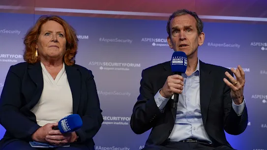 A woman with red hair in a dark blazer and a man in a dark suit speaking on a panel at the Aspen Security Forum. The man is holding a microphone and gesturing while speaking, while the woman sits beside him holding her own microphone. The background is a blue wall with the Aspen Institute and Aspen Security Forum logos.