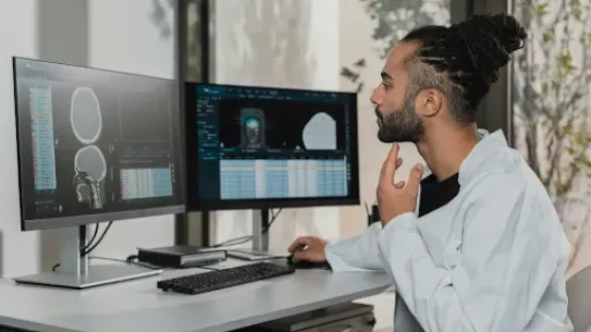 A male medical professional or researcher with hair in a bun and wearing a white lab coat sits at a desk, thoughtfully analyzing medical brain scans and data on two large computer monitors in a bright, modern office.