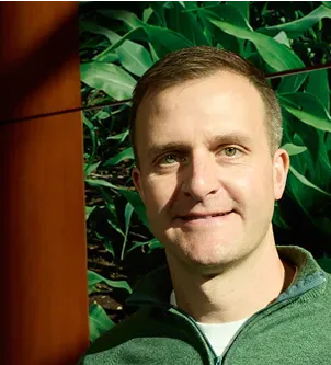 Close-up portrait of a man in a green quarter-zip sweater, smiling in front of a background of lush green plants and a wooden pillar.
