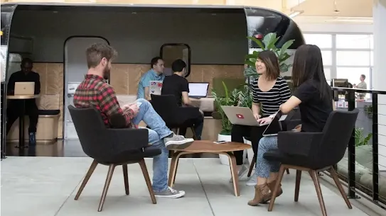 A group of colleagues working in a modern, open-concept office space featuring a large, black indoor trailer used as a meeting or lounge area. In the foreground, three people sit in dark gray upholstered chairs around a small wooden table, using laptops and a smartphone. Behind them, other employees are seated at desks and inside the trailer, creating a collaborative and dynamic professional environment.