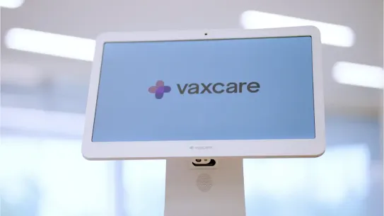 A white VaxCare touchscreen kiosk displaying the company logo on a blue background.