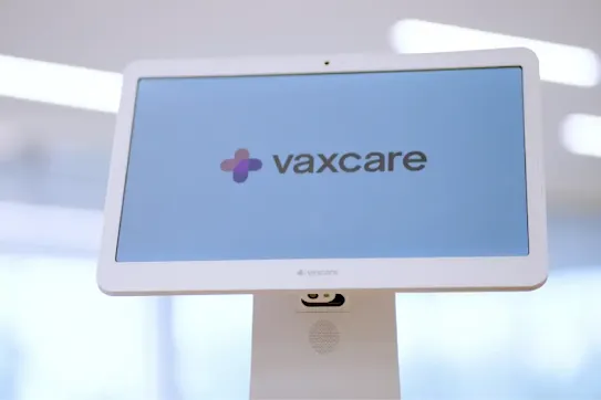 A white VaxCare touchscreen kiosk displaying the company logo on a blue background.
