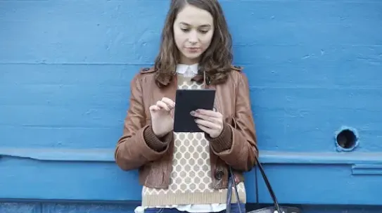 A woman in a brown leather jacket and patterned sweater stands against a blue wall while using a black tablet.