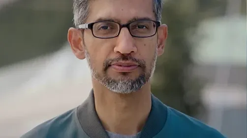 A professional headshot of Sundar Pichai, CEO of Google and Alphabet. He is looking directly at the camera, wearing black-rimmed glasses, a gray t-shirt, and a dark teal bomber jacket. The background is softly blurred, focusing attention on his neutral and composed expression.