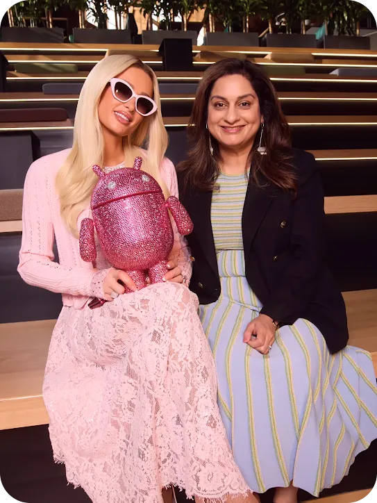 Paris Hilton holding a bedazzled pink Android mascot, sitting on wooden bleachers next to a smiling woman in a striped dress and black blazer.
