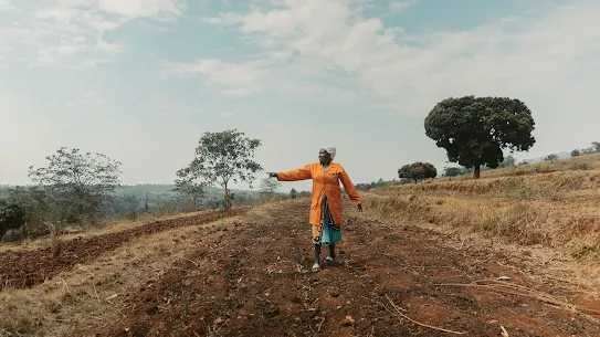 A woman in an orange jacket standing on a tilled dirt path in a rural agricultural field and pointing into the distance under a bright sky.