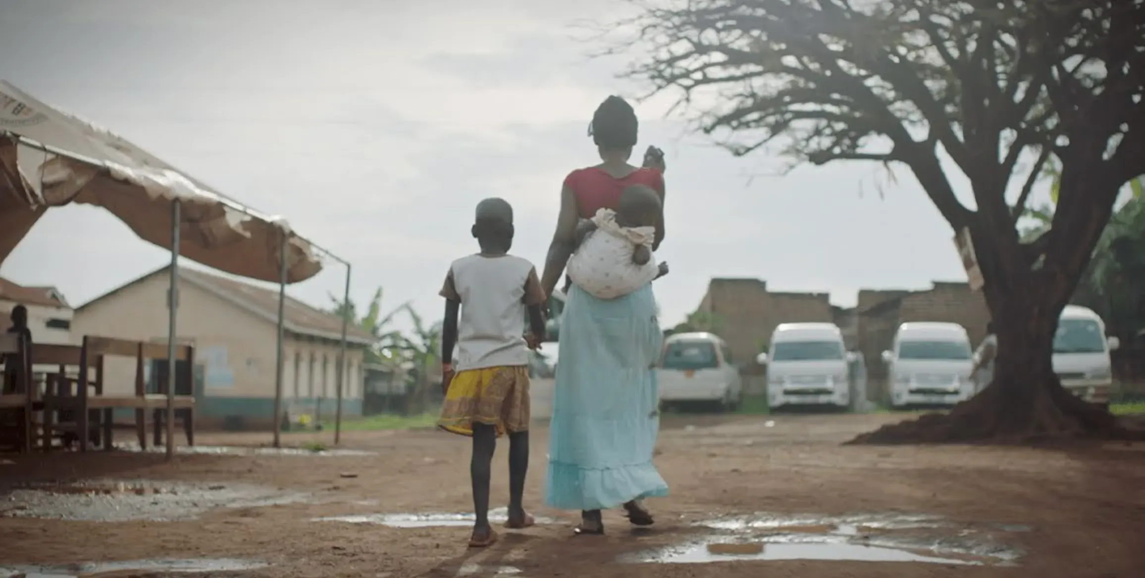 A woman carries her baby on her back and holds her daughter’s hands as they walk away along a dirty road.