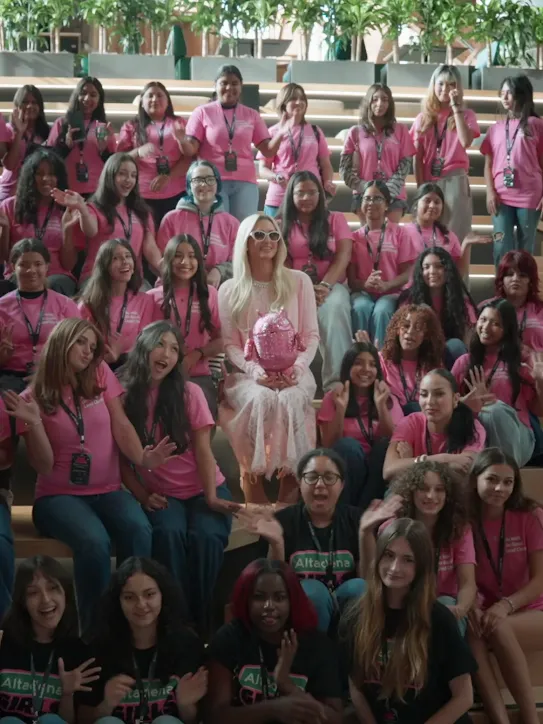 Paris Hilton wearing white sunglasses and holding a bedazzled pink Android mascot, sitting on wooden bleachers surrounded by a large group of smiling and waving girls in pink and black shirts.