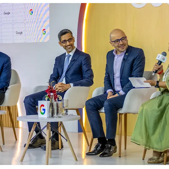 Four panelists sit on a stage during an AI Summit event. From left to right: a man in a blue blazer, Sundar Pichai in a dark suit, a man in a navy blazer with glasses, and a woman in a green traditional dress holding a microphone and speaking. Two small white tables with Google-branded cubes and water bottles are in front of them.