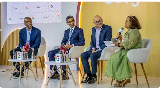 Four panelists sit on a stage during an AI Summit event. From left to right: a man in a blue blazer, Sundar Pichai in a dark suit, a man in a navy blazer with glasses, and a woman in a green traditional dress holding a microphone and speaking. Two small white tables with Google-branded cubes and water bottles are in front of them.